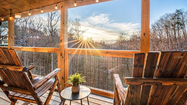 Eine Holzterrasse mit zwei Stühlen und einem kleinen Tisch, mit Blick auf eine bewaldete Landschaft unter blauem Himmel.
