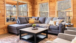 Ein gemütliches Blockhäuschen-Interieur mit einer Eckcouch, einem Couchtisch und großen Fenstern mit Blick auf den umliegenden Wald.