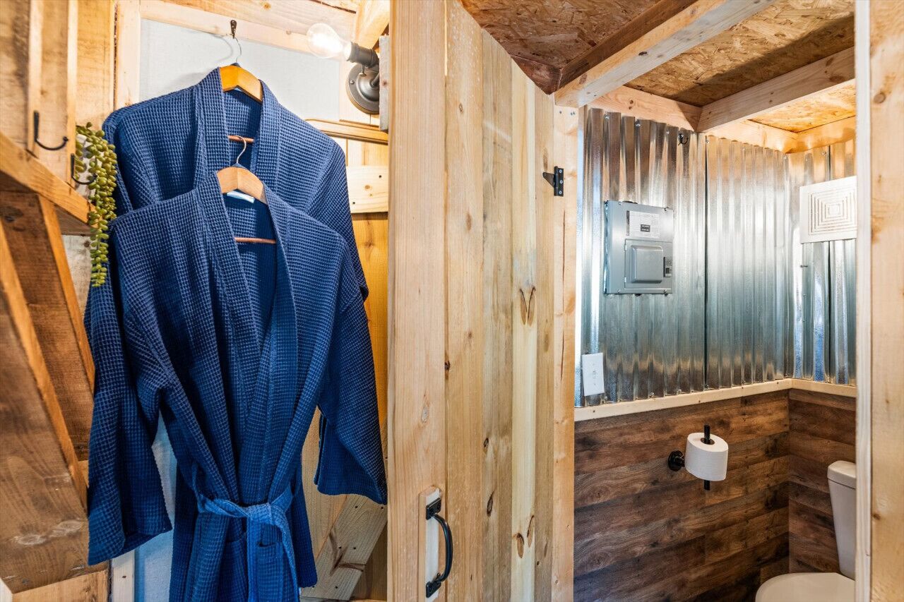 Een rustieke houten badkamer met een houten badjas hangend aan een rek, een toilet en een raam met metalen frame.