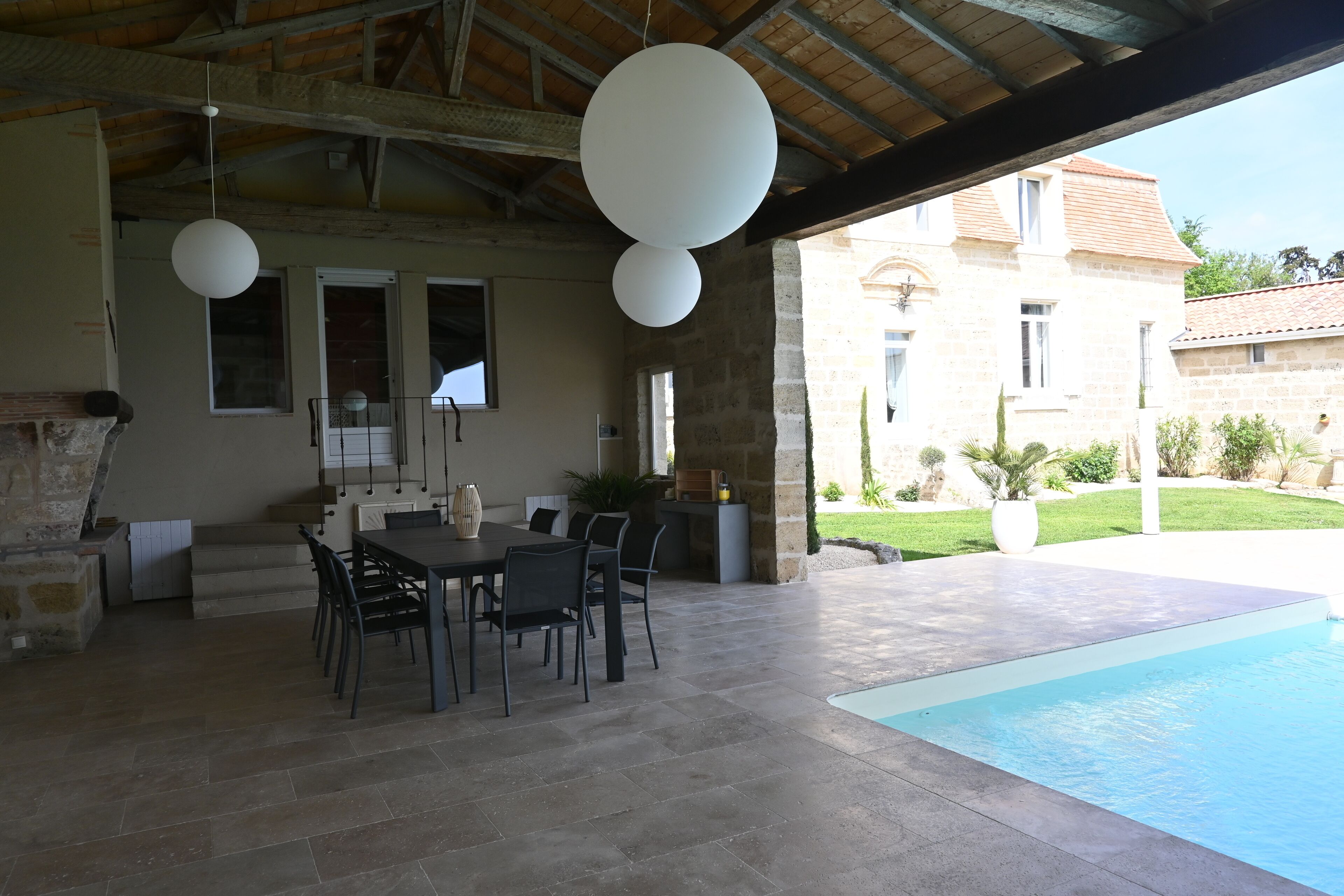 An outdoor patio with a pool, stone flooring, a dining table with chairs, and a stone fireplace.