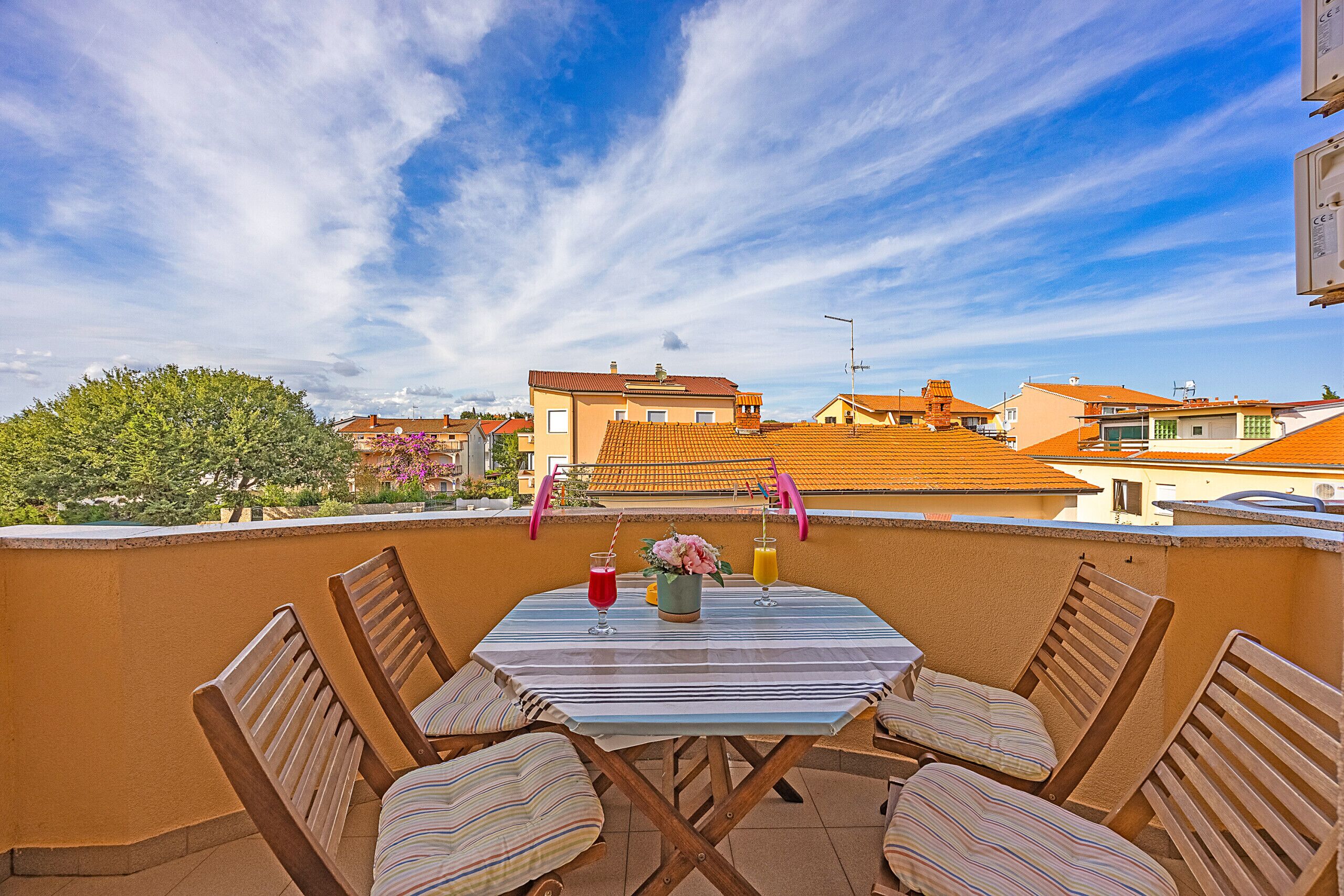 A rooftop terrace with a table set for two, featuring striped cushions and a vase with flowers.