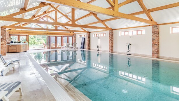 An indoor swimming pool with wooden beams, a glass wall, and a patio area with lounge chairs. - Cedar Barn - UKC5406 (Banham)