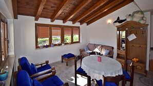 A cozy room with a wooden ceiling, a round table with a lace tablecloth, blue upholstered chairs, a sofa, and a wooden cabinet with hooks.