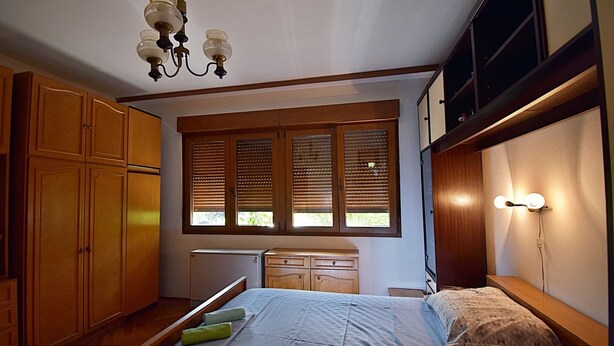 A bedroom with a bed, wooden wardrobe, a window with blinds, and a ceiling light fixture.