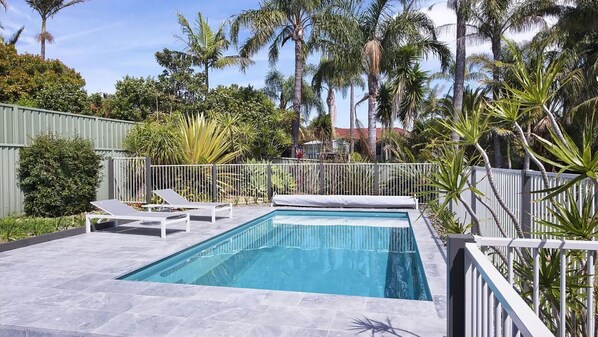 A rectangular swimming pool with a tiled deck, surrounded by a white fence and palm trees.