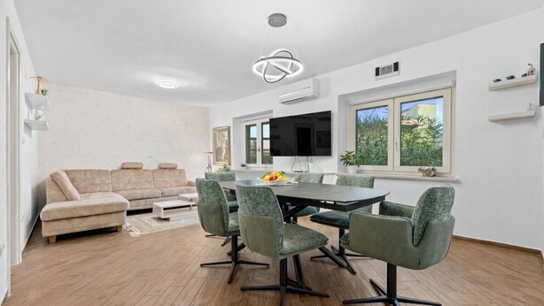A modern living room with a beige sofa, a dining table with chairs, a flat-screen TV, and a large window with a view of greenery. - Apartment Pineta close to the Beach (Medulin)