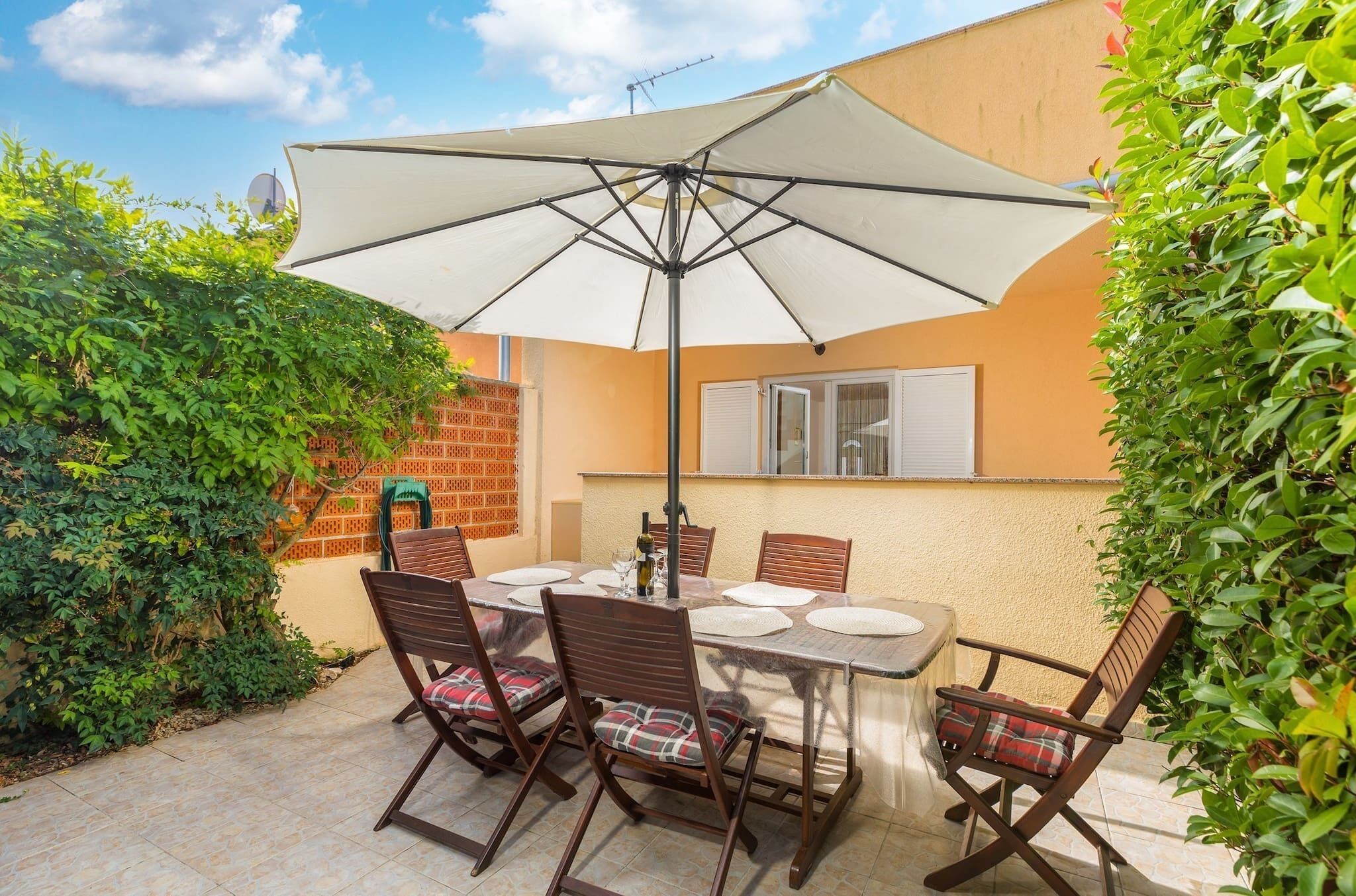 Um pátio com mesa e cadeiras sob um grande guarda-sol, cercado por vegetação e uma parede de tijolos.