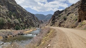 A dirt road alongside a river with rocky mountains in the background.