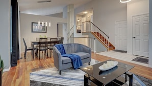 A modern living room with a grey sofa, dining table, and chairs. A staircase leads to an upper level.