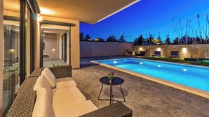 Un espace extérieur avec une piscine, un canapé et des fauteuils, une petite table agrémentée d’une plante, et une vue sur le ciel au crépuscule.