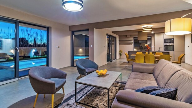 Un salon moderne avec un canapé, des fauteuils, une table basse et un coin repas avec des chaises jaunes. On peut apercevoir une piscine à travers les portes vitrées.