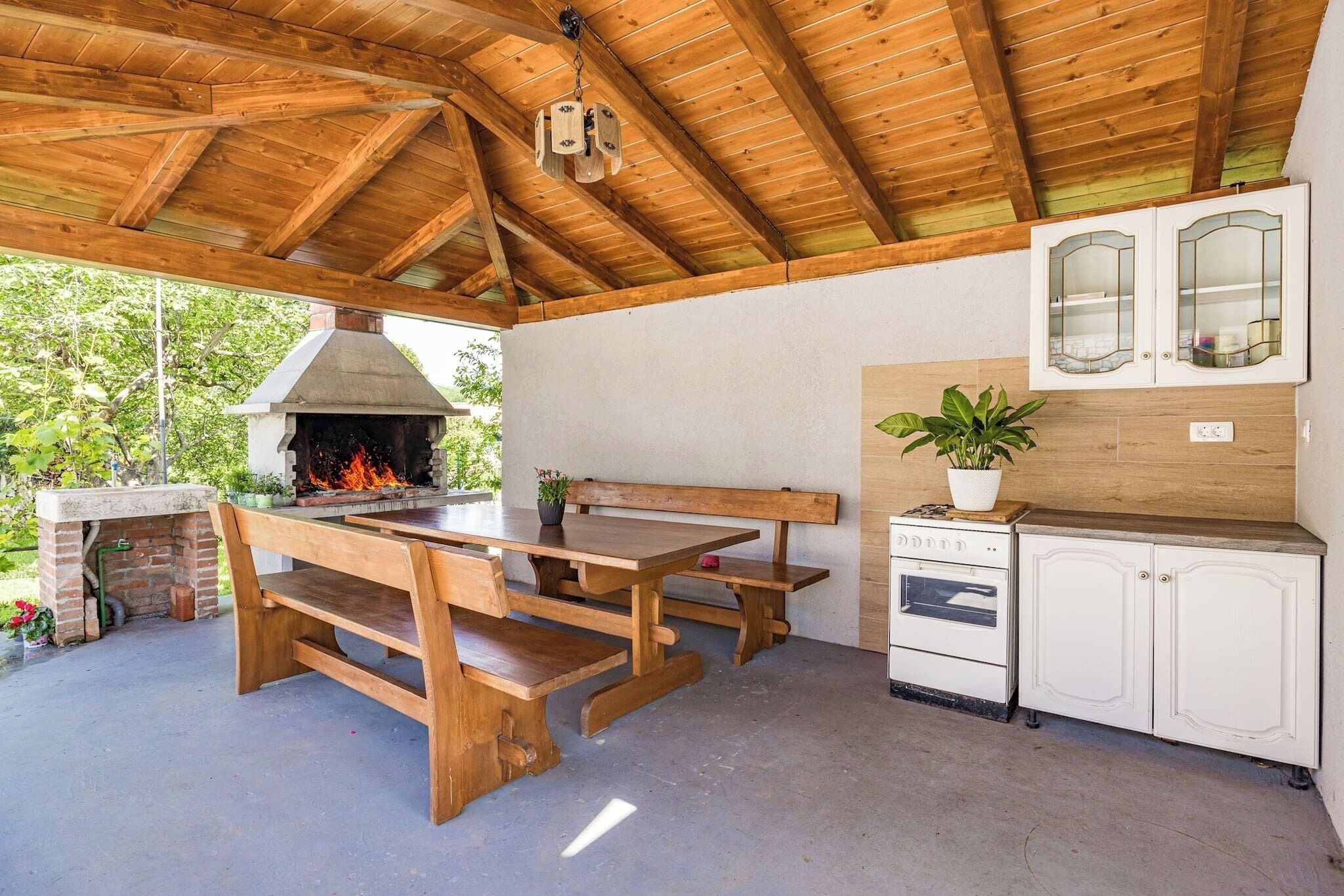 An outdoor kitchen area with a wooden table and benches, a brick oven, and a white cabinet with glass doors.