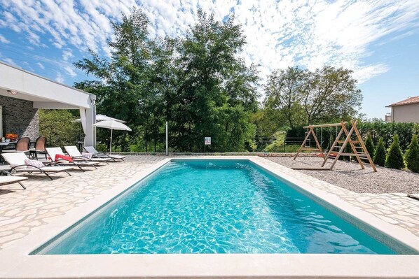 Una piscina rectangular con agua azul cristalina, rodeada de sillas de descanso y un conjunto de columpios de madera.