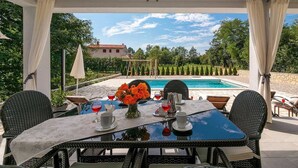 A covered outdoor dining area with a table set for two, overlooking a swimming pool and a landscaped garden. - Villa Sofia with Private Pool (Santalezi)