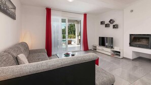 A modern living room with a grey sofa, a flat-screen TV, a fireplace, and a glass door leading to an outdoor area with a pool. - Villa Sofia with Private Pool (Santalezi)