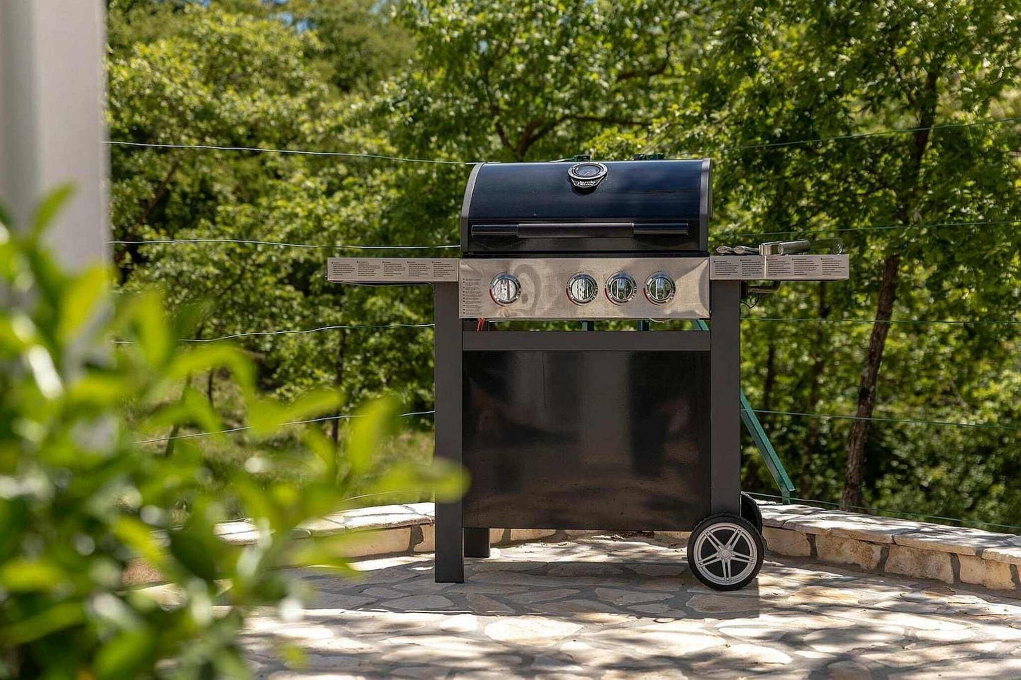 Un barbecue portatif doté de quatre brûleurs, d’un couvercle et de roulettes pour faciliter son déplacement, installé sur une terrasse en pierre avec un décor d’arbres en arrière-plan.