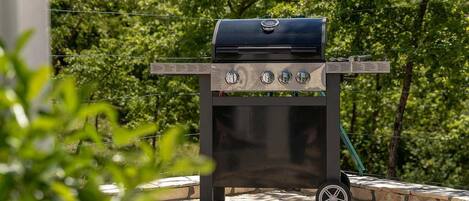 Un barbecue portatif doté de quatre brûleurs, d’un couvercle et de roulettes pour faciliter son déplacement, installé sur une terrasse en pierre avec un décor d’arbres en arrière-plan.