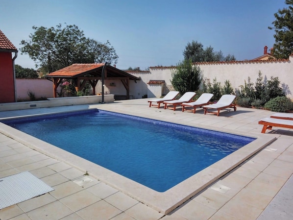 Une piscine rectangulaire dont le bord est recouvert de carreaux, entourée d’une terrasse également carrelée. On peut voir quatre chaises longues alignées parallèlement à la piscine, et une petite structure couverte avec un toit carrelé se trouve à l’arrière-plan.