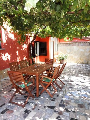 Une table et des chaises en bois sont disposées sur une terrasse carrelée, avec un mur rouge et un mur en pierre en arrière-plan.