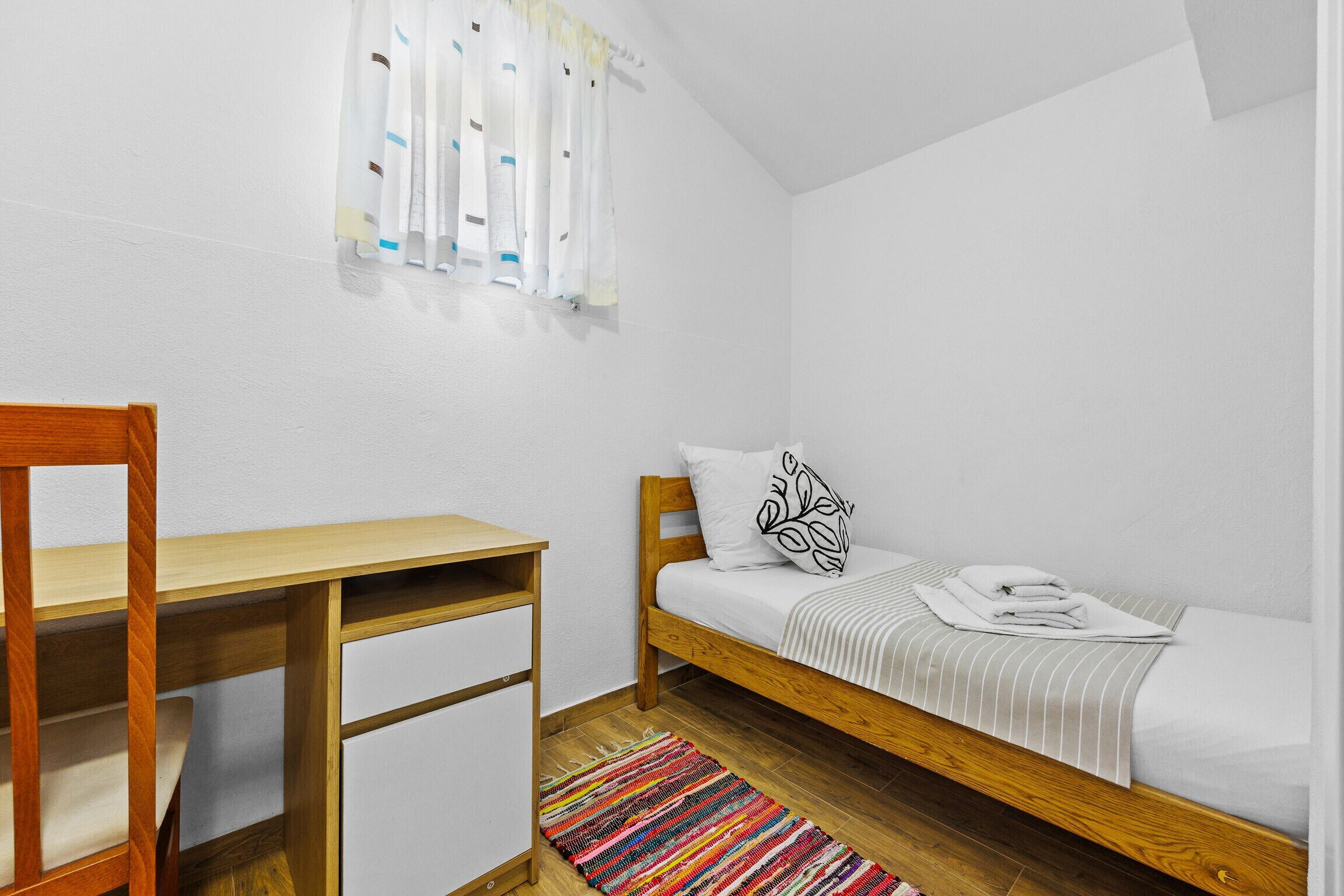 Ein kleiner, ordentlicher Raum mit einem hölzernen Bett, einem Schreibtisch und einem farbenfrohen StreifenTeppich.