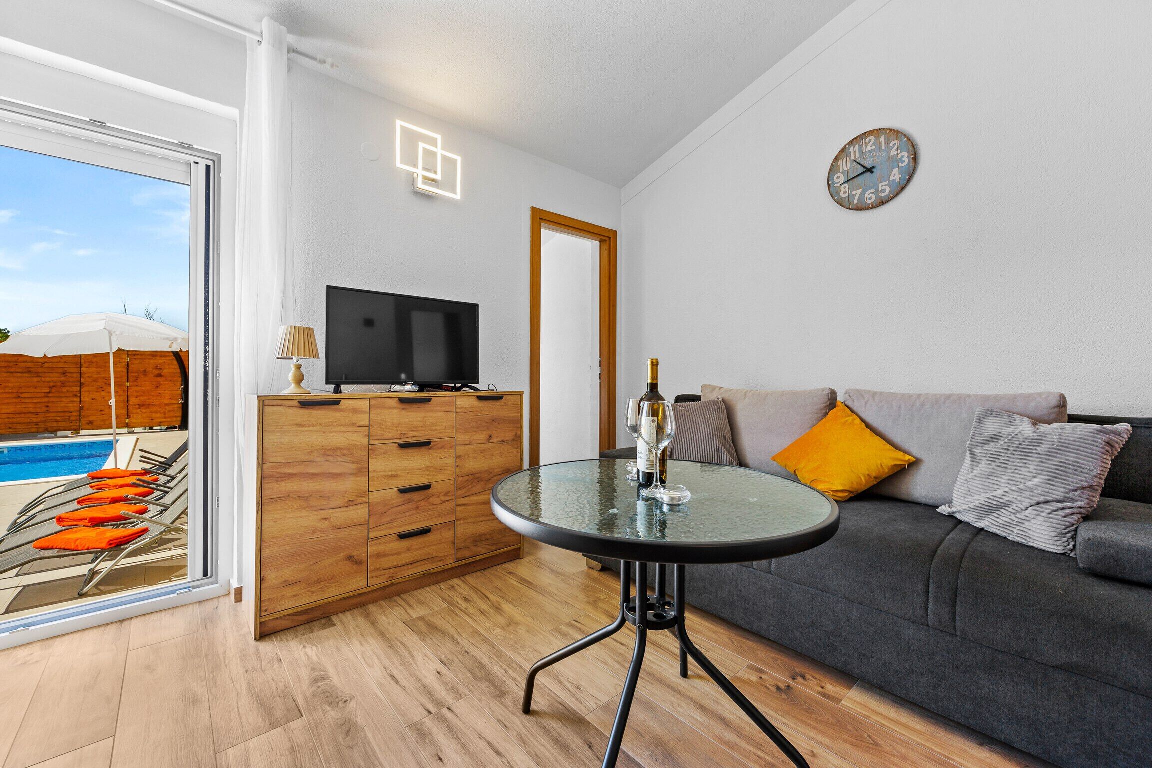 A modern living room with a glass table, a sofa with cushions, a television on a wooden cabinet, and a clock on the wall.
