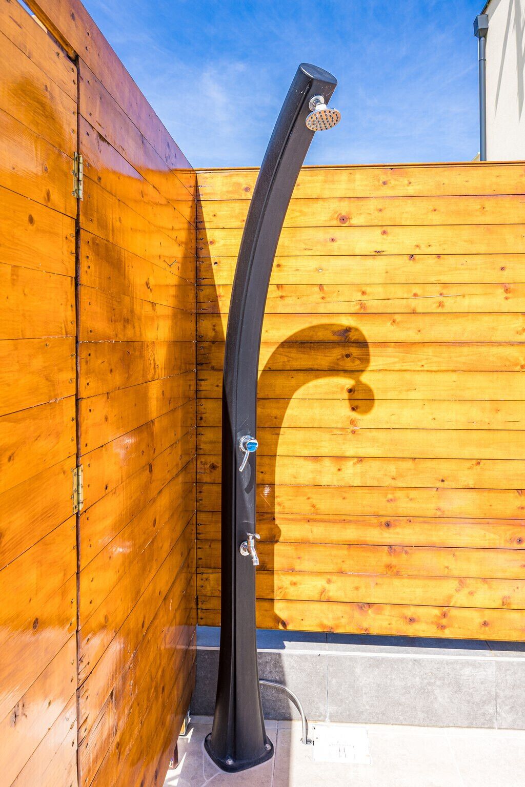 Une douche extérieure noire au design incurvé, fixée sur une base en béton, adossée à une clôture en bois.
