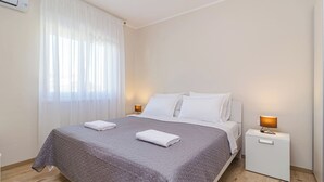 Un lit bien fait, recouvert d’une couette grise, avec des oreillers blancs et deux serviettes blanches pliées. Il y a une table de chevet avec une lampe et un petit tiroir. La pièce a des murs blancs et une fenêtre avec des rideaux légers.