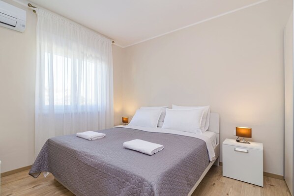 Ein ordentlich bezogenes Bett mit grauer Decke, weißen Kissen und zwei gefalteten weißen Handtüchern. Ein Nachttisch mit Lampe und kleiner Schublade steht daneben. Die Wände sind weiß und es gibt ein Fenster mit durchsichtigen Vorhängen.