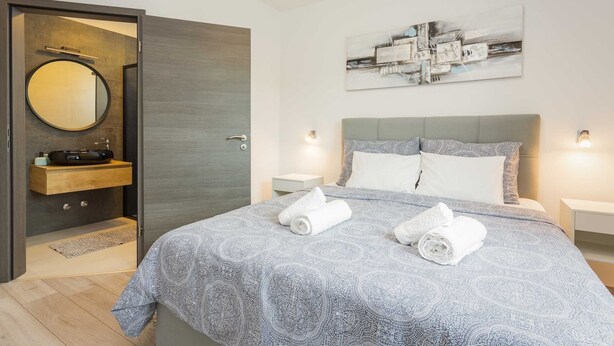 A modern bedroom with a large bed, a bedside table, a nightstand, a wall-mounted artwork, and a bathroom with a mirror and sink. - Holiday Home Greta with Private Pool (Salakovci)