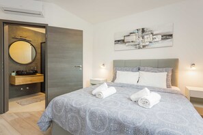 A modern bedroom with a large bed, a bedside table, a nightstand, a wall-mounted artwork, and a bathroom with a mirror and sink. - Holiday Home Greta with Private Pool (Salakovci)