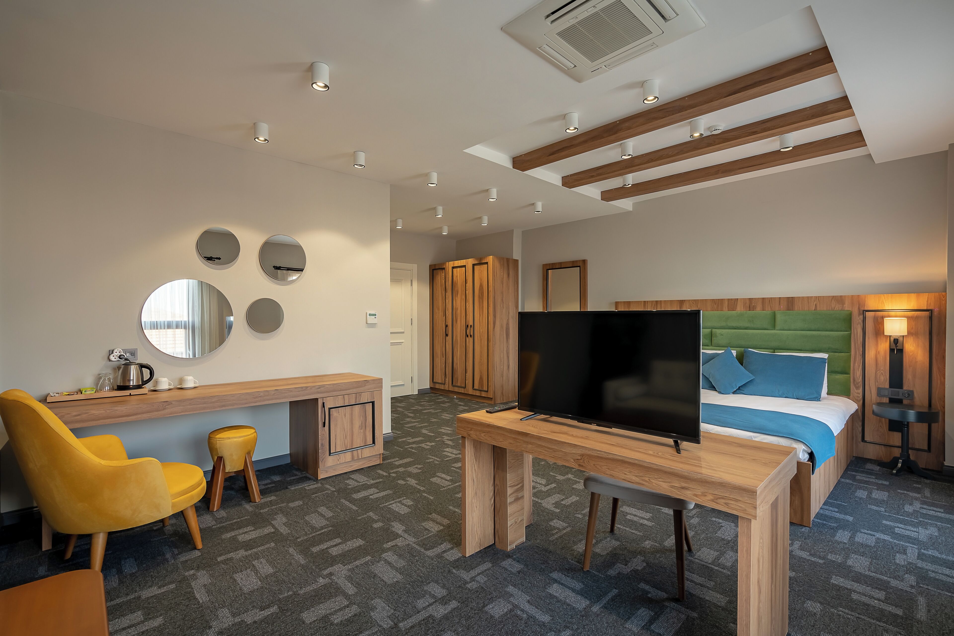 A hotel room with a wooden desk, a TV, a bed with green headboard, and a yellow chair.