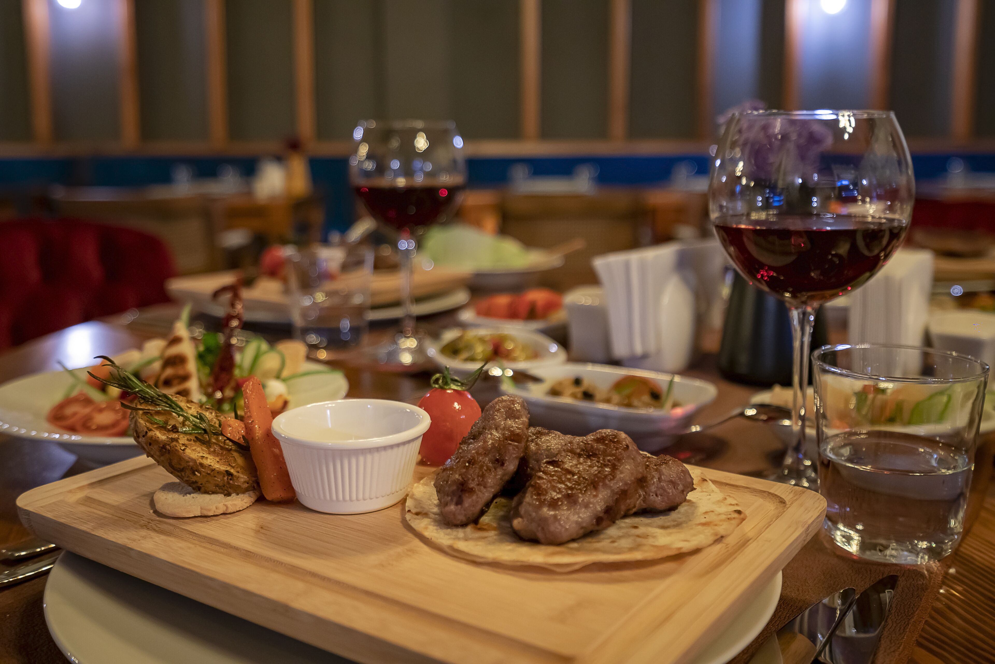 A dining table with a variety of dishes, including grilled meat, vegetables, and a small bowl of sauce. There are also glasses of red wine and a glass of water.