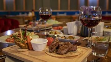 A dining table with a variety of dishes, including grilled meat, vegetables, and a small bowl of sauce. There are also glasses of red wine and a glass of water.