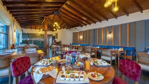 Restaurante com vigas de madeira no teto, mesas redondas com cadeiras estofadas em vermelho, e diversos pratos sobre as mesas.