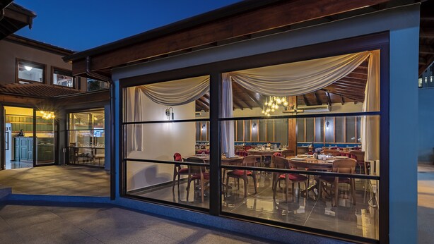 Um restaurante com grandes janelas, área de refeições com mesas e cadeiras, e cozinha visível.