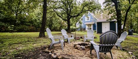 Un patio trasero con un brasero, sillas Adirondack blancas y una casa azul.