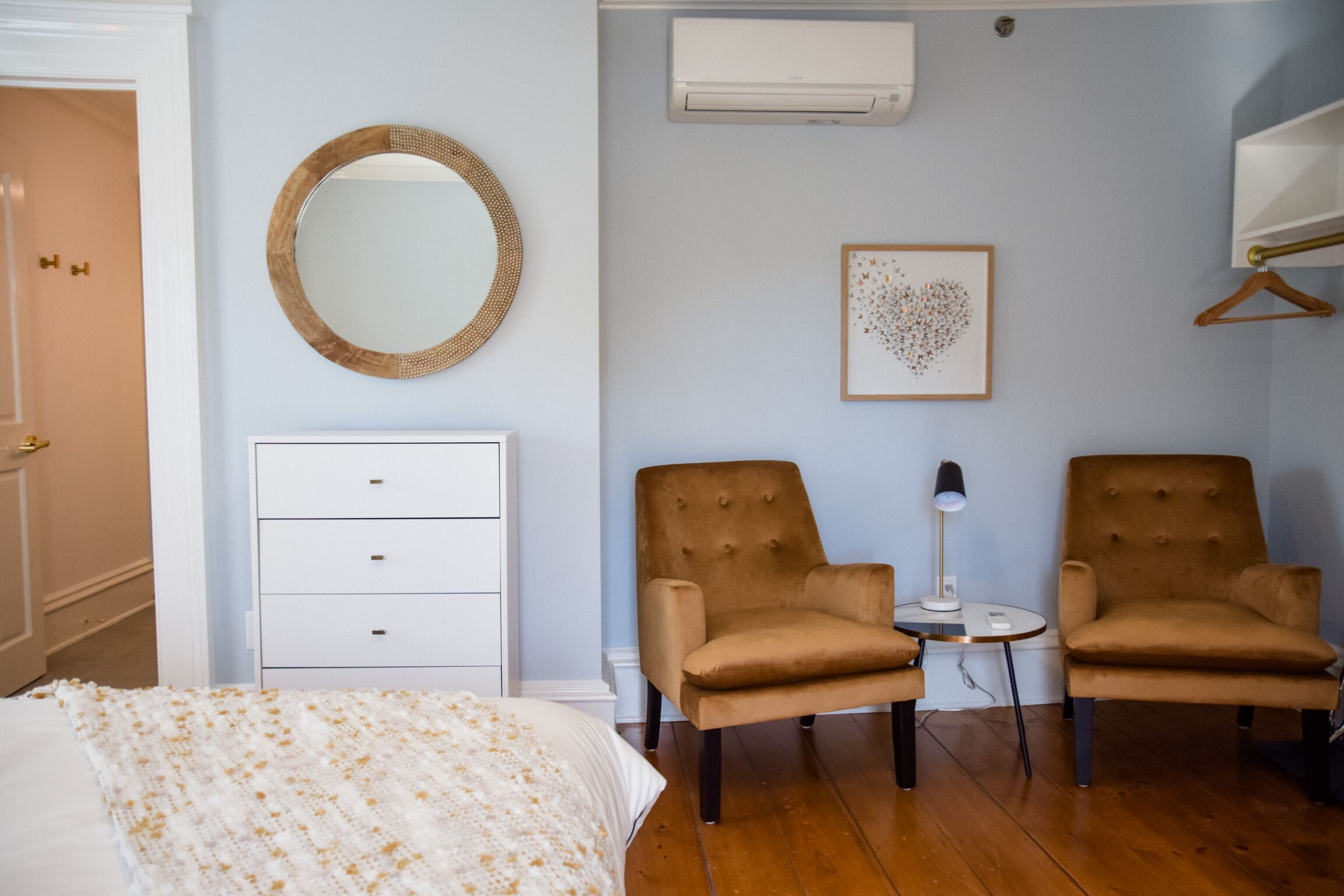Un dormitorio con una cama, un aparador, dos sillones, una mesita con una lámpara, un espejo montado en la pared, un aire acondicionado y un cuadro enmarcado.