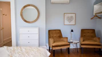 Ein Schlafzimmer mit einem Bett, einem Schminktisch, zwei Sesseln, einem kleinen Tisch mit Lampe, einem wandmontierten Spiegel, einer Klimaanlage und einem gerahmten Bild an der Wand.