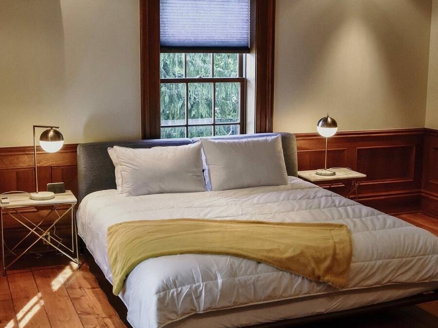 A neatly made bed with white bedding and a yellow throw blanket, positioned in a room with wooden floors and a large window with blinds.