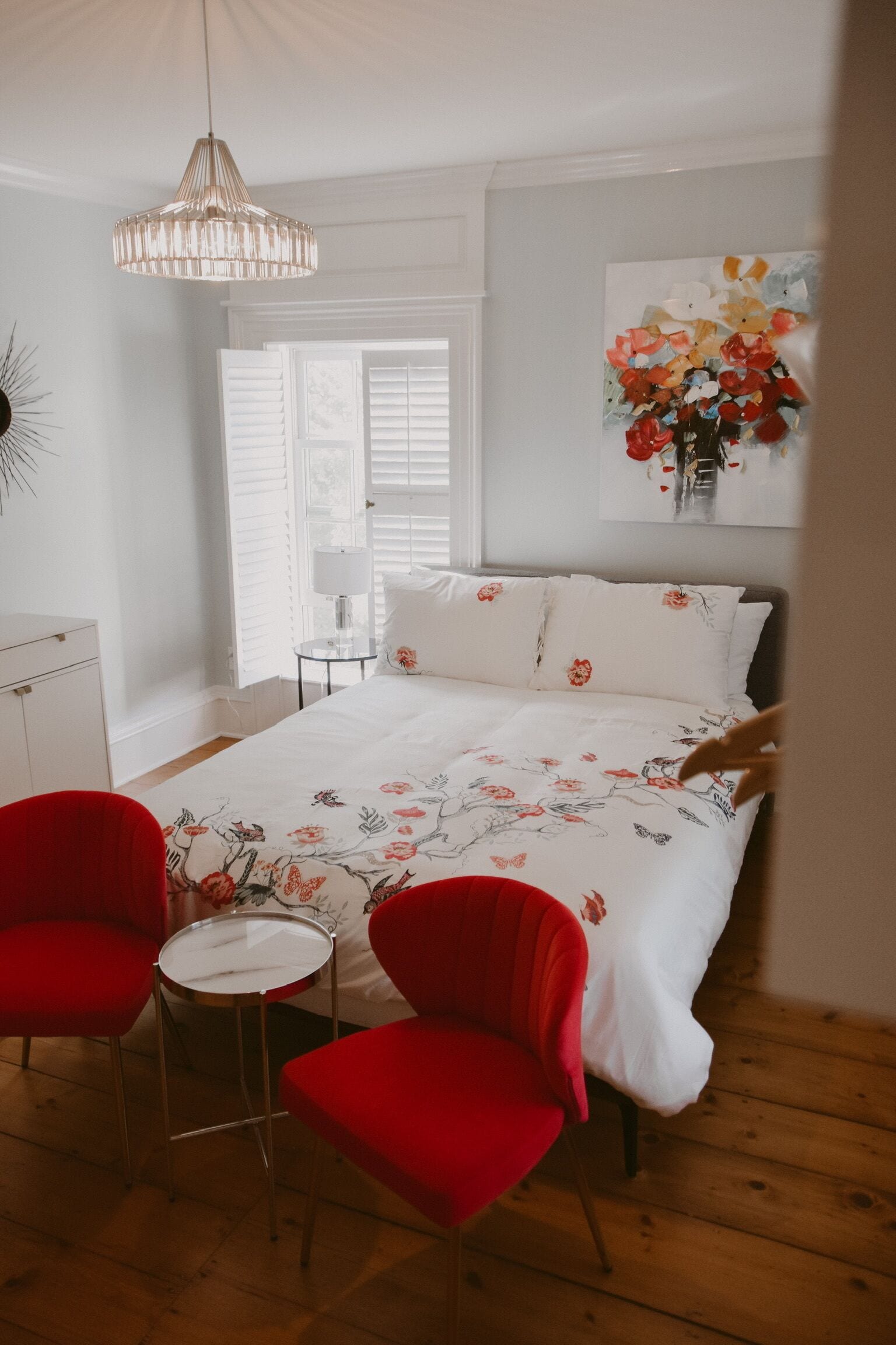 Ein Schlafzimmer mit einem Bett, zwei roten Sesseln, einem kleinen Tisch, einer Pendelleuchte und einem Fenster mit Jalousien.