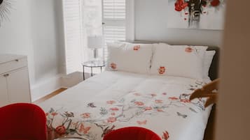 Ein Schlafzimmer mit einem Bett, zwei roten Sesseln, einem kleinen Tisch, einer Pendelleuchte und einem Fenster mit Jalousien.