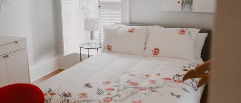 Un dormitorio con una cama, dos sillas rojas, una mesita, una lámpara colgante y una ventana con persianas.