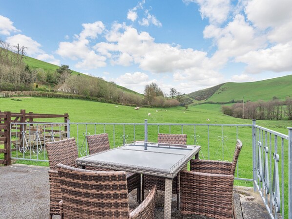 Un espace extérieur aménagé avec des meubles en osier et une table, offrant une vue sur un champ verdoyant où paissent des moutons.