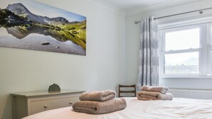 Ein Schlafzimmer mit einem Bett, einem Sessel, einem Nachttisch, einem Gemälde und einem Fenster mit Vorhängen.