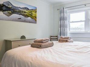 Een slaapkamer met een bed, een stoel, een nachtkastje, een schilderij en een raam met gordijnen.