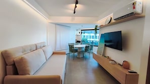A modern living room with a sofa, a dining table with chairs, a flat-screen TV mounted on the wall, and a small shelf with a vase and remote control.