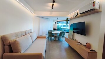 Una sala de estar moderna con sofá, una mesa de comedor con sillas, un televisor de pantalla plana montado en la pared y una pequeña estantería con un florero y un control remoto.