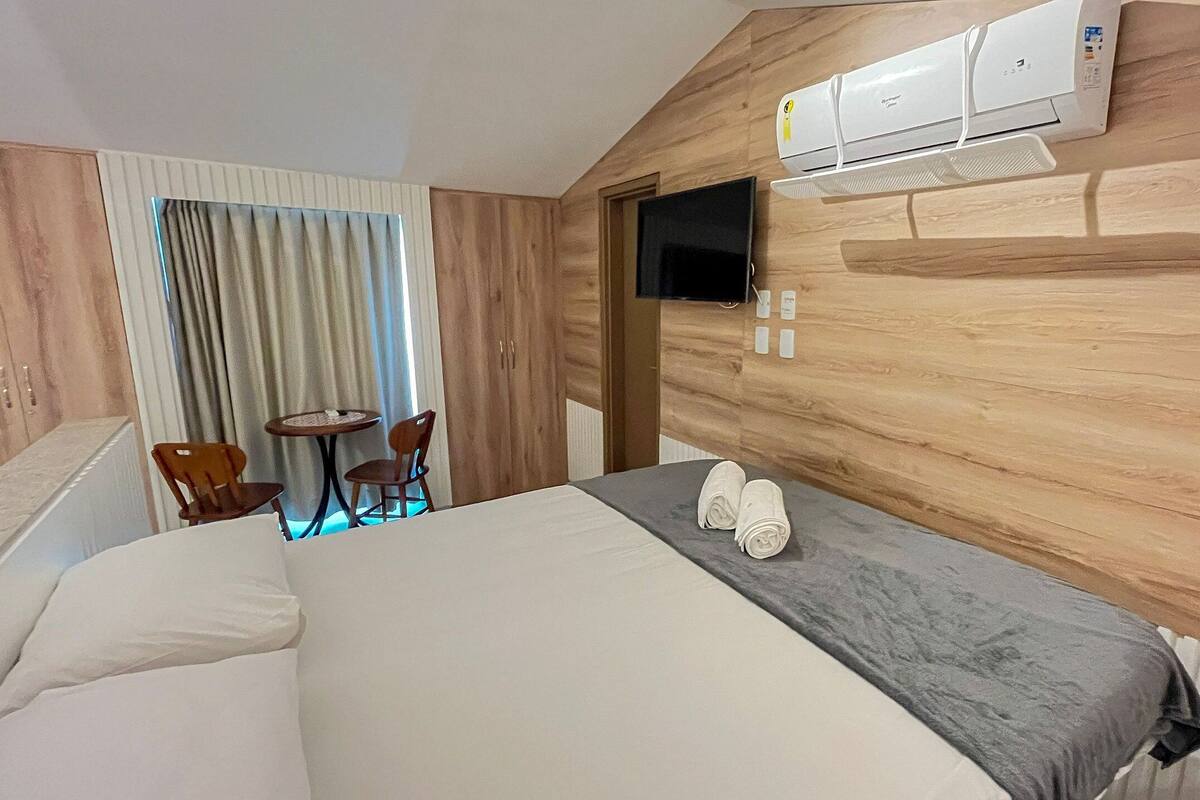 A hotel room with a bed, a TV, an air conditioner, a small table with chairs, and a view through a window.