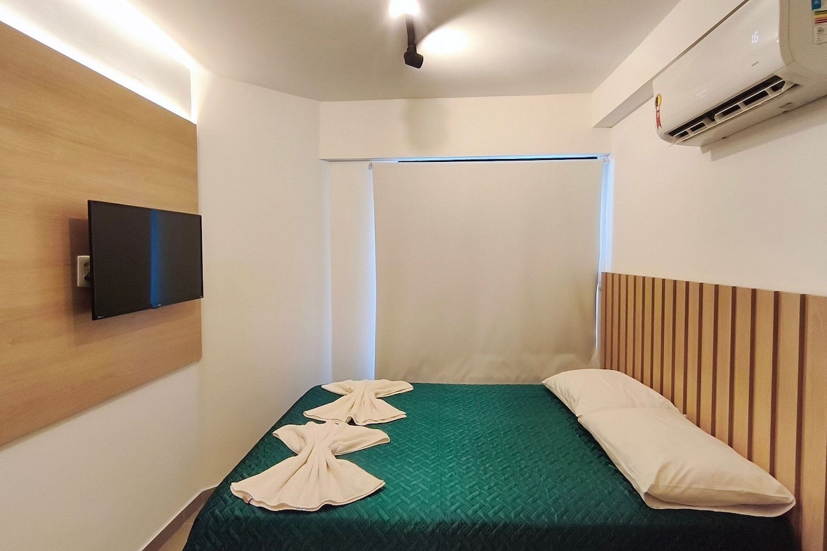 A small, neatly arranged room with a bed, a flat-screen TV mounted on the wall, and an air conditioning unit.