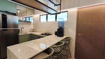 Una cocina moderna con un refrigerador de acero inoxidable, un horno empotrado, una estufa a gas y una cafetera. Hay dos taburetes de mimbre y una ventana sobre la encimera.